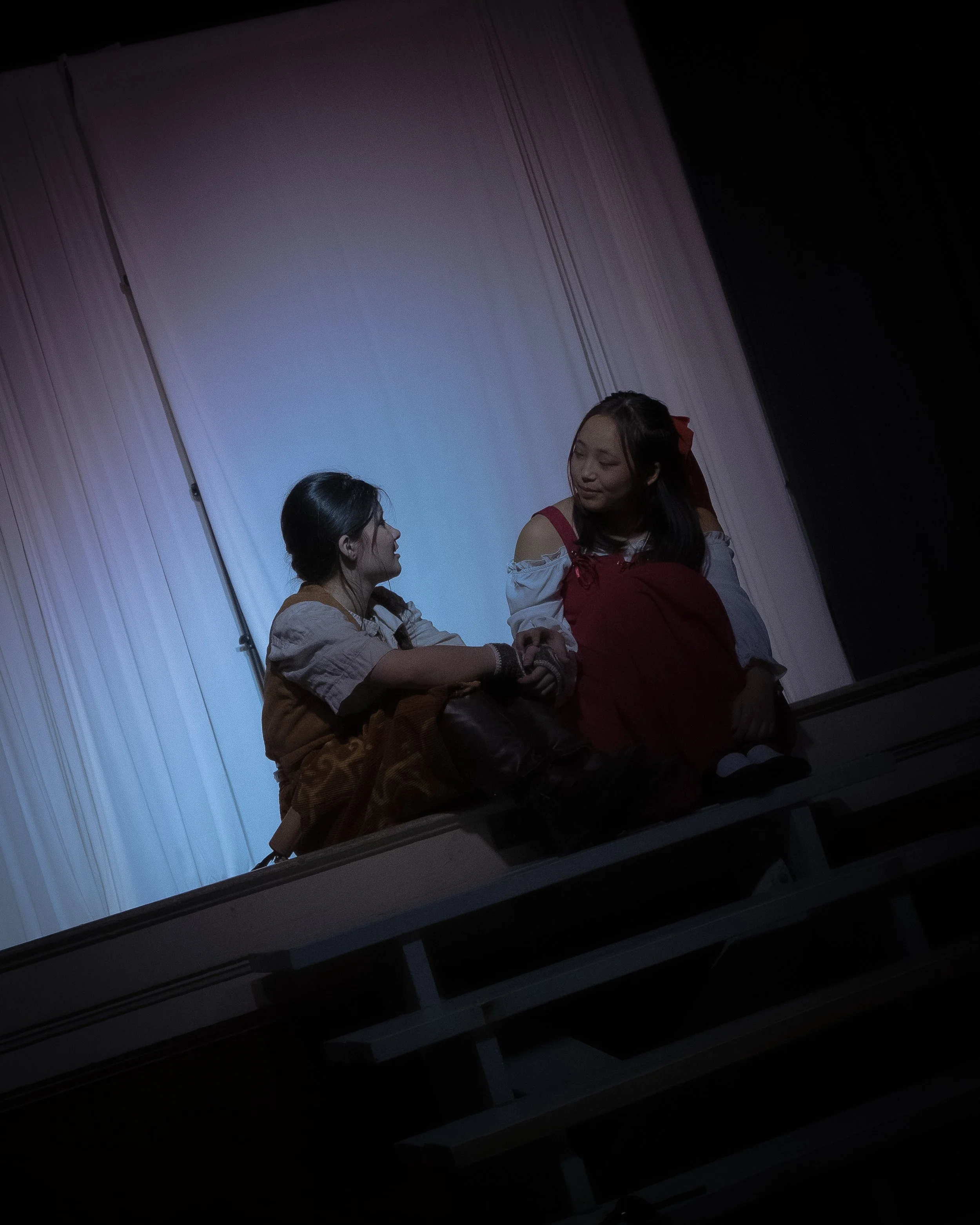 Two young women in costume sitting on a stage, holding hands and looking at each other, with a blue-white curtain in the background.