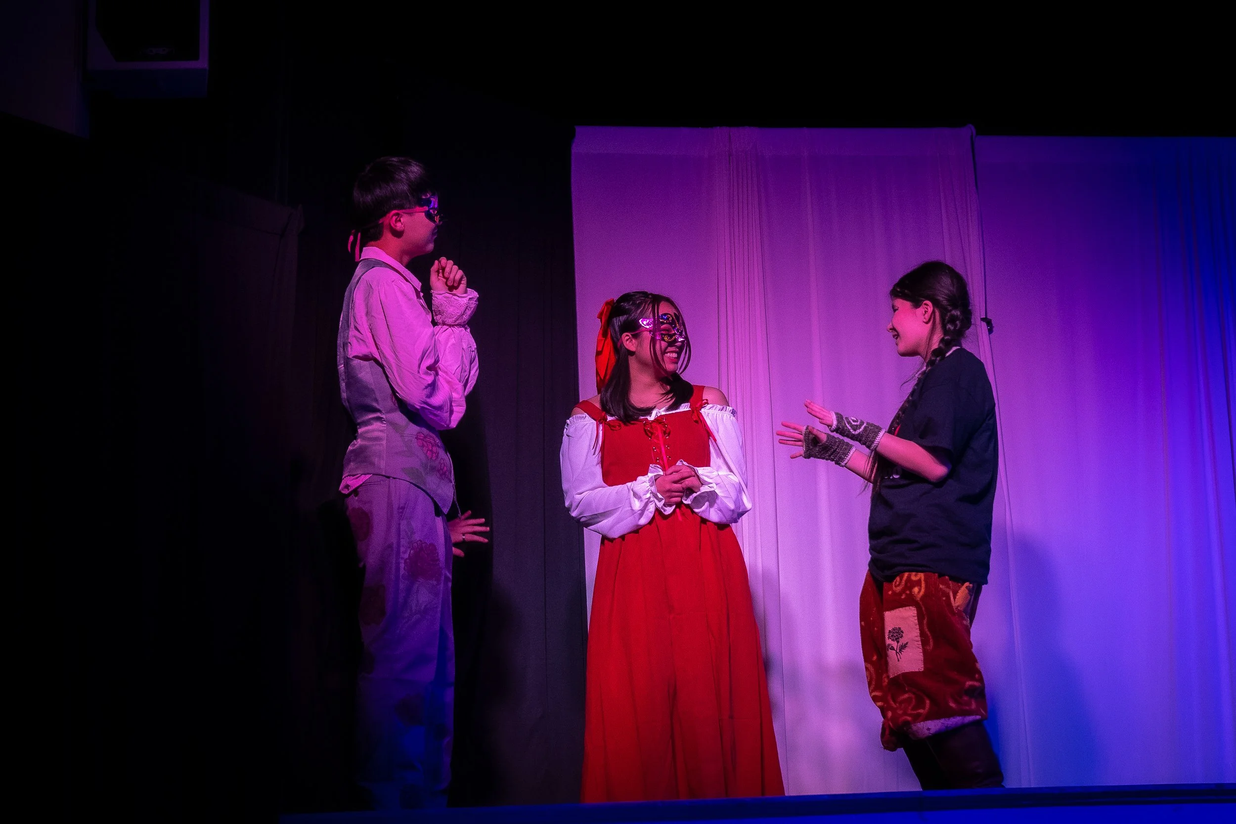 Three children performing in a theater, with purple and blue stage lighting. Two girls and one boy are dressed in colorful costumes, engaged in conversation as part of a play.