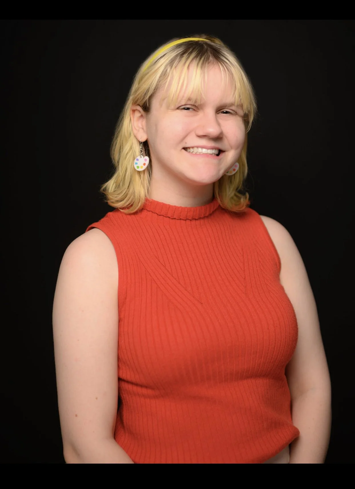 A young woman with blonde hair wearing a red sleeveless top smiling against a black background. She has paint palette earrings and a subtle smile.
