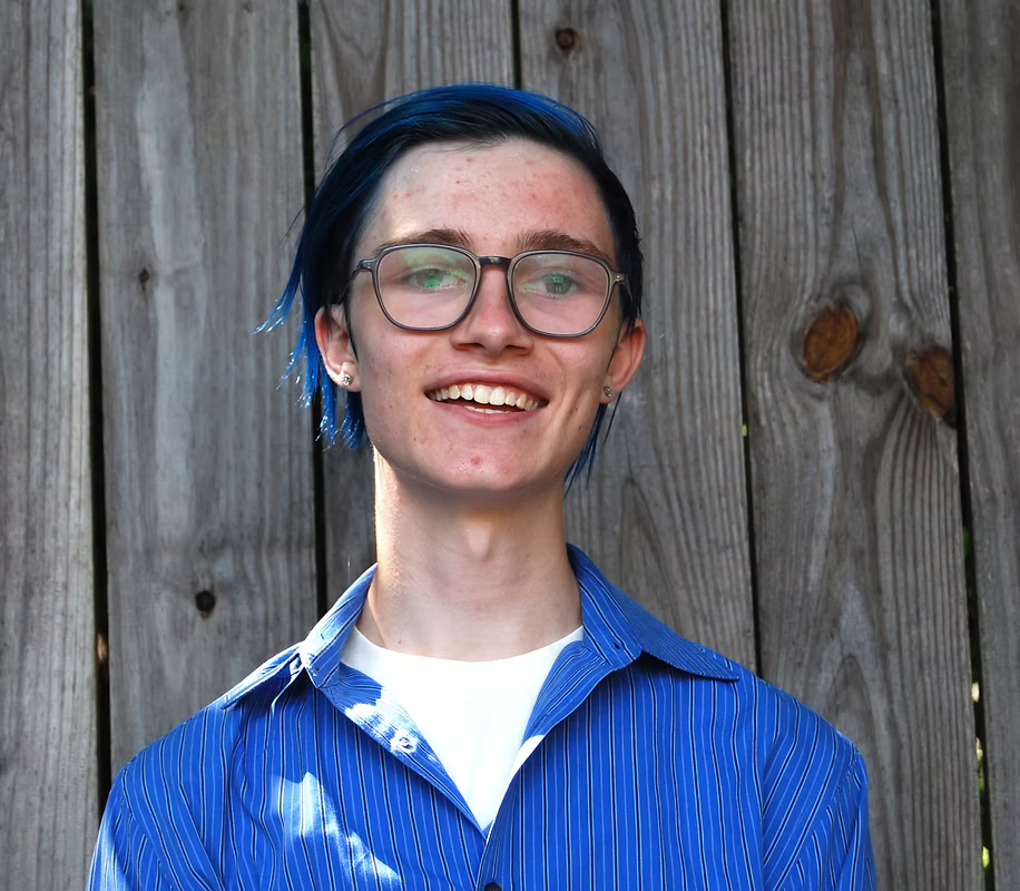Young person with blue hair and glasses smiling, wearing a blue striped shirt, standing in front of a wooden fence.