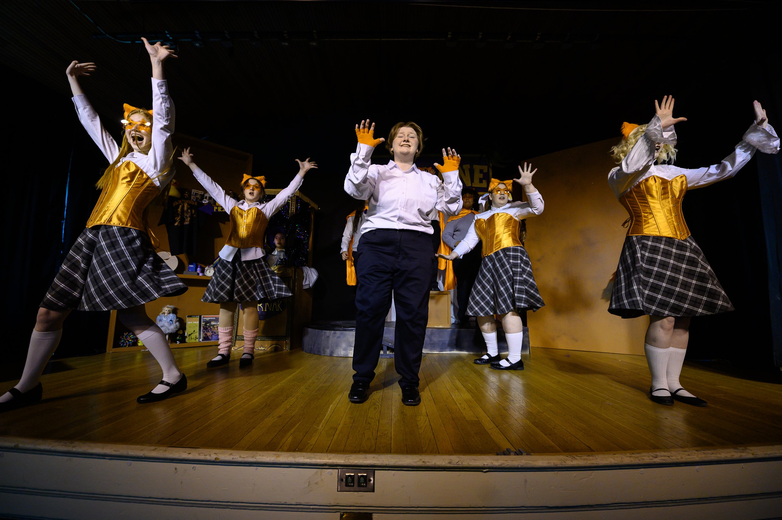Children performing a stage play with costumes that include plaid skirts, orange vests, and cat headbands, with a person in the center smiling and gesturing. This is a photo from The Roguish Players' production of Ride The Cyclone