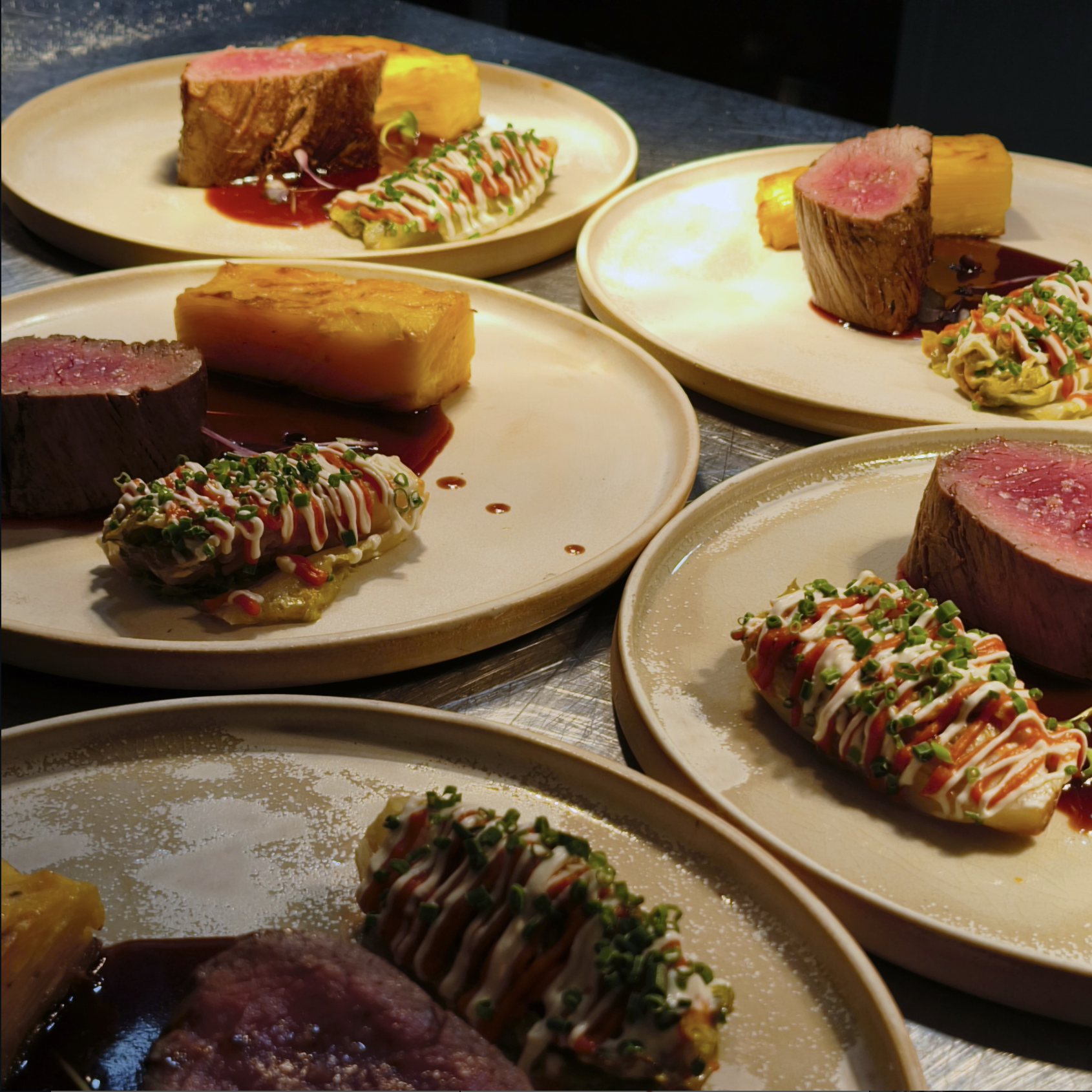 Mehrere Teller mit rotem und pinkem Fleisch, Kartoffelgratin, asiatischem Sushi, und einer Saucenähnlichen Zubereitung, alles an einem dunklen Tisch.
