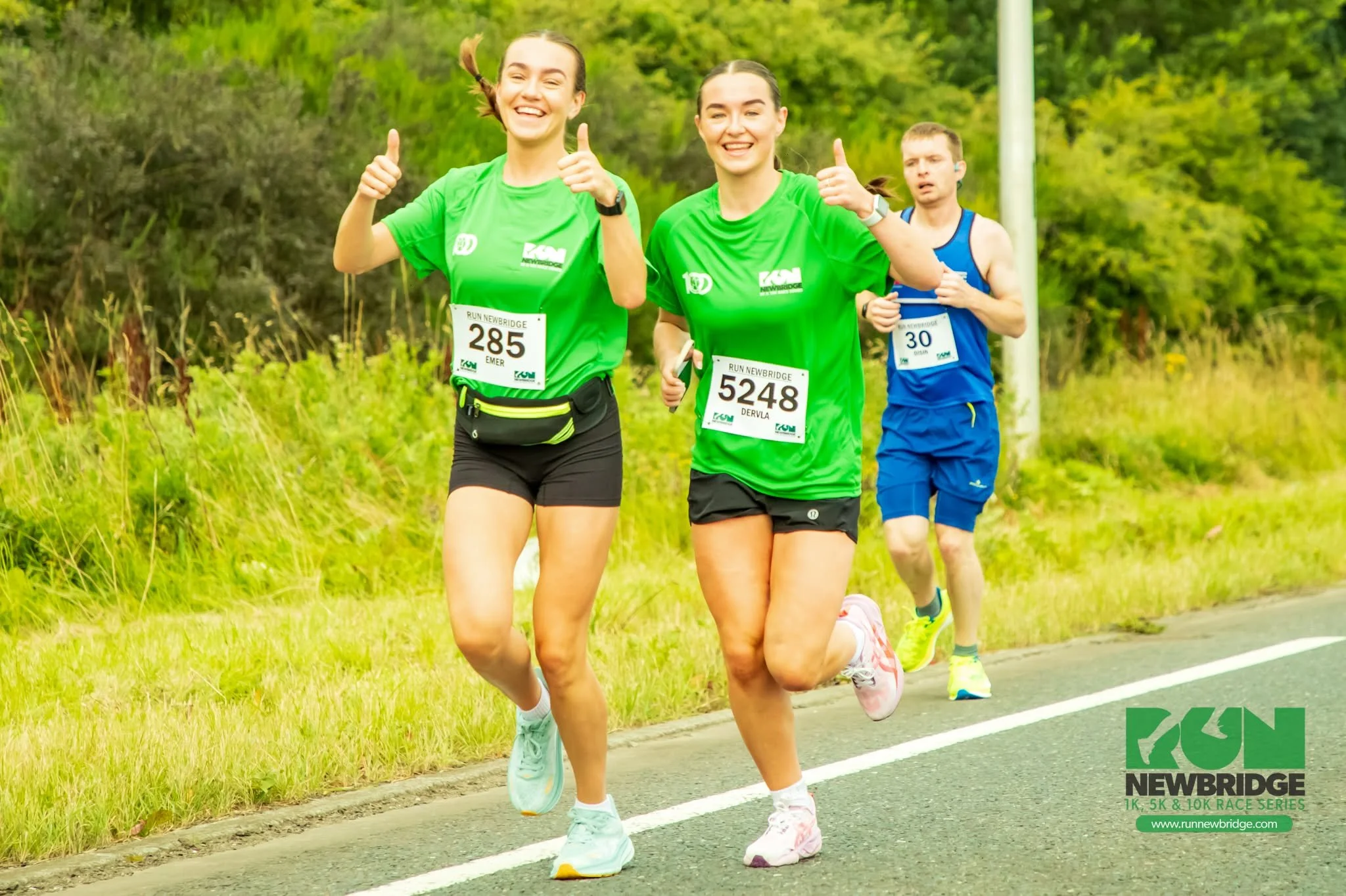 Three runners participating in a race, running along a paved path with greenery in the background, two women smiling and giving thumbs-up, and a man running behind them.