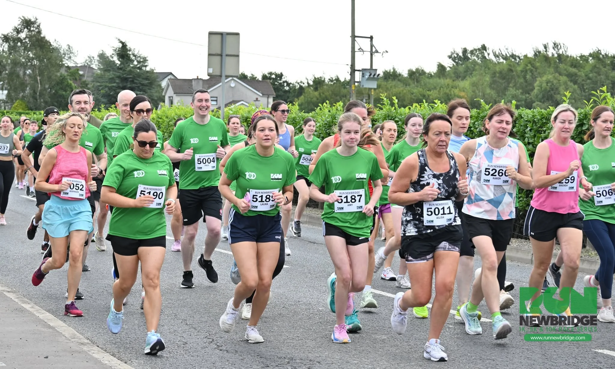 A group of people participating in a running event, wearing athletic clothes and bib numbers, running on a street with greenery and houses in the background.