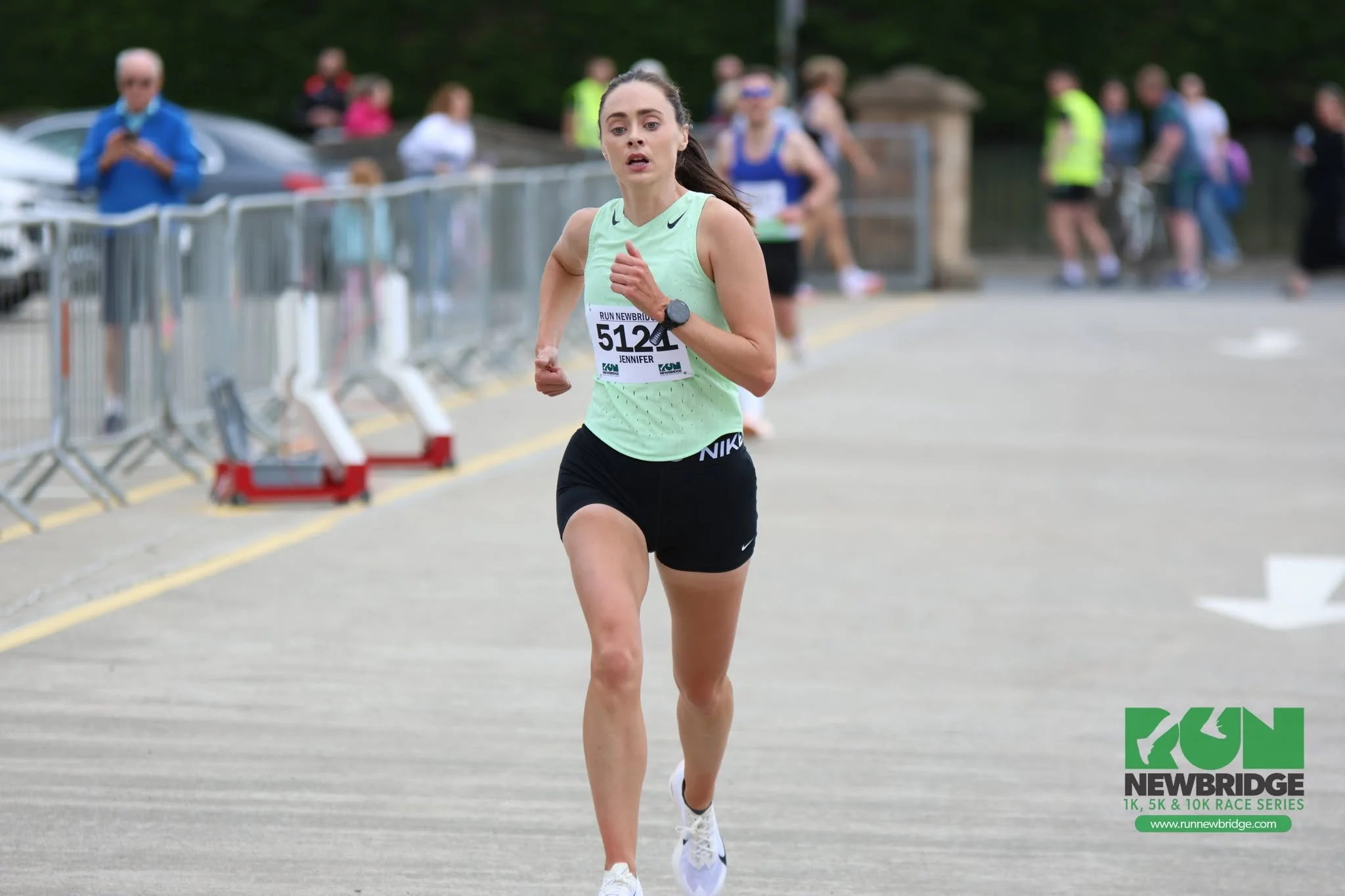 A woman in a light green tank top and black shorts running in a race, wearing a race bib with the name Jennifer and number 5121, with spectators and race officials in the background.
