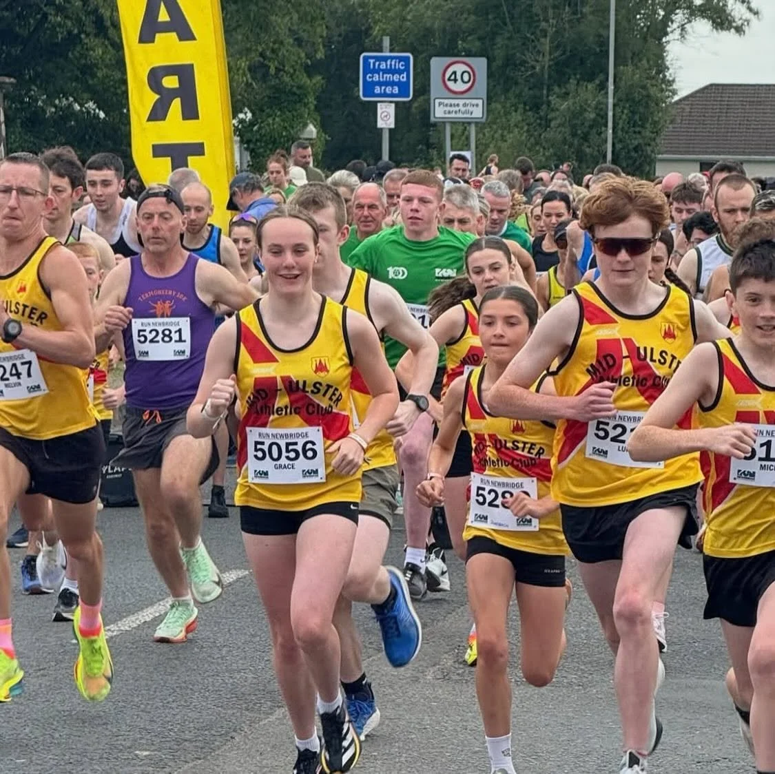 A group of runners participating in a race, wearing athletic gear and bib numbers, on a street in a crowd with signs and green trees in the background.