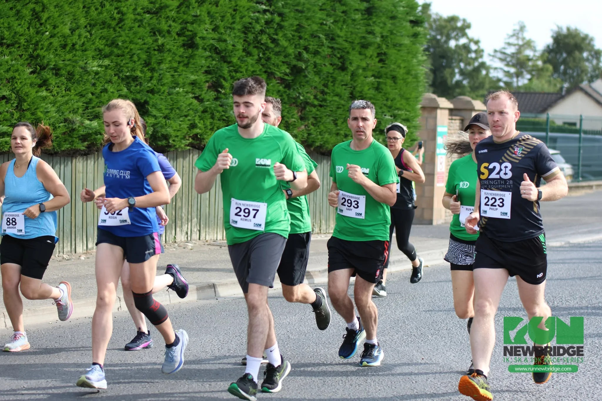 Group of runners participating in a race, wearing athletic clothing with race bibs, running on a street with greenery and houses in the background.