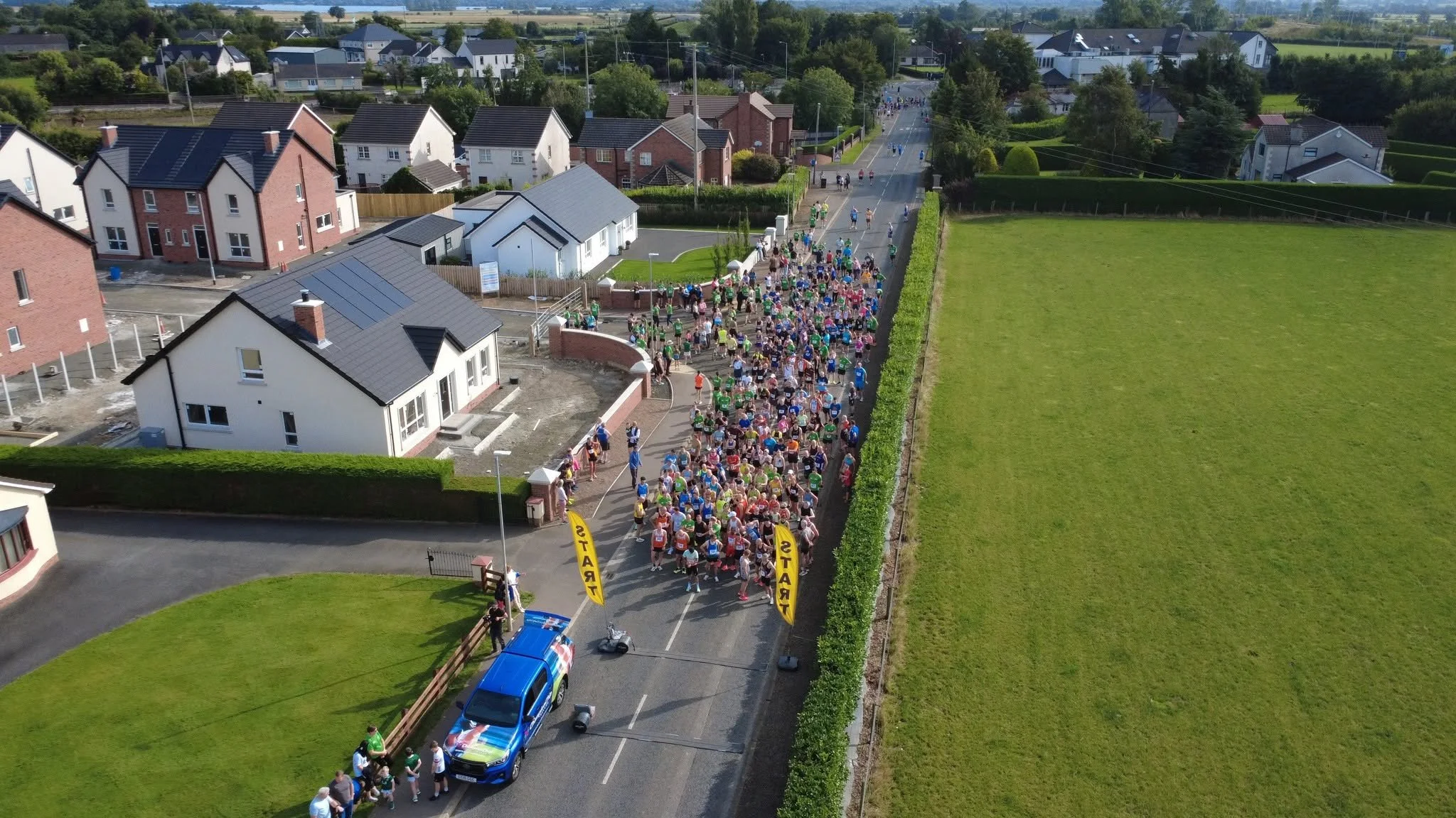 Aerial view of a large group of runners participating in a race or marathon on a suburban street, with houses and green fields on either side.