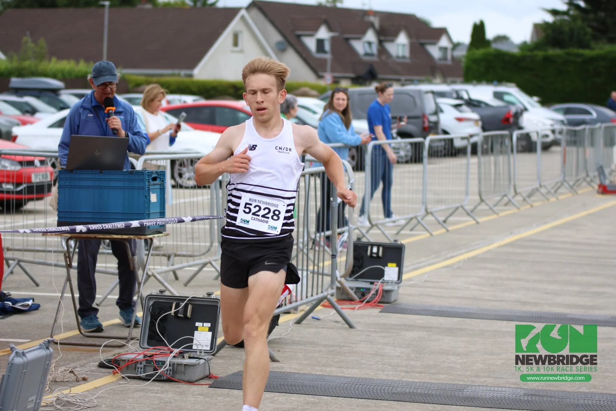 Male runner crossing the finish line at a race event, with a race number 5228, near a parking lot and event staff with electronic equipment.