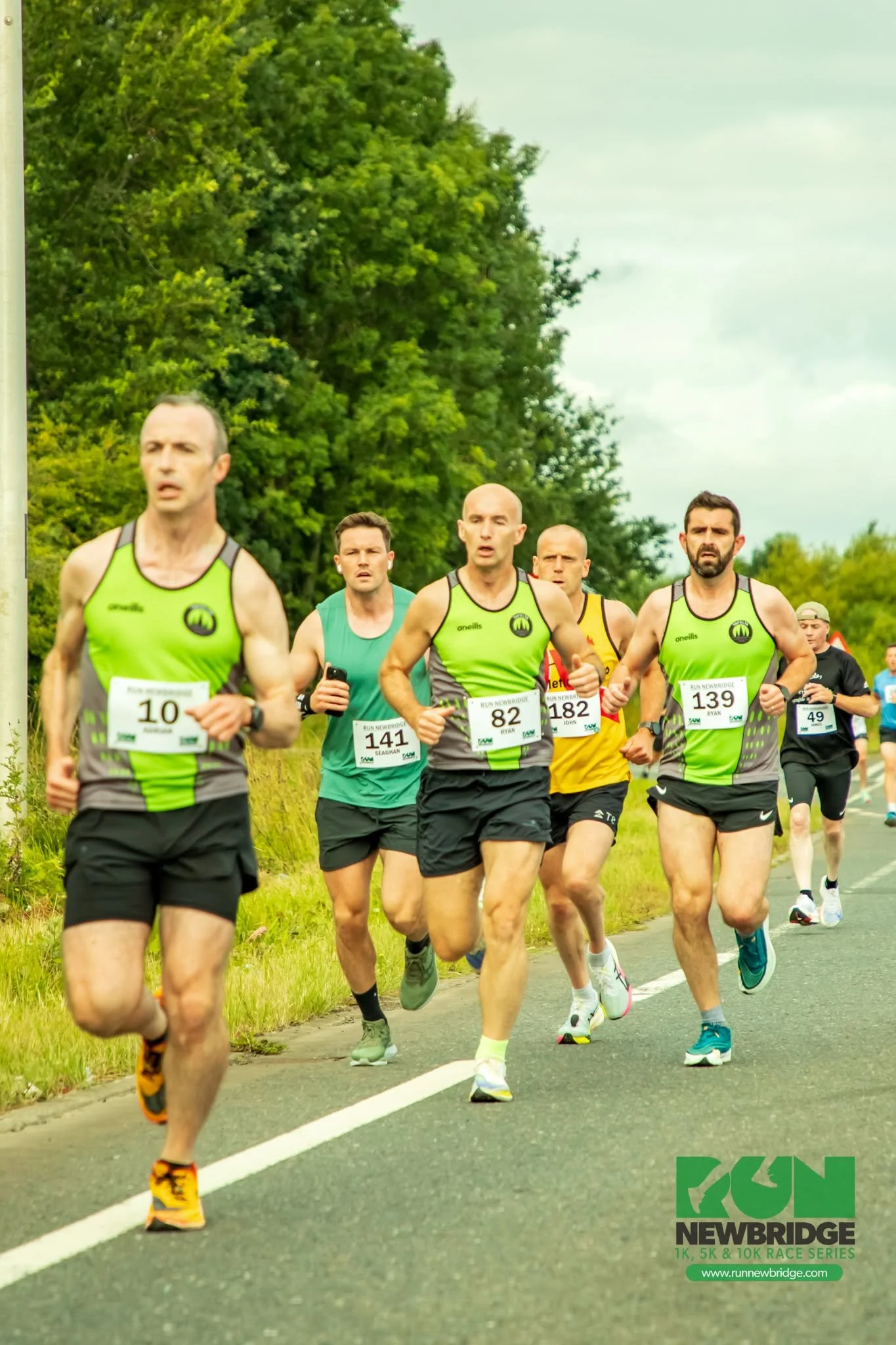 A group of male runners participating in a race on a road surrounded by greenery, with some of them wearing race bibs and athletic clothing.