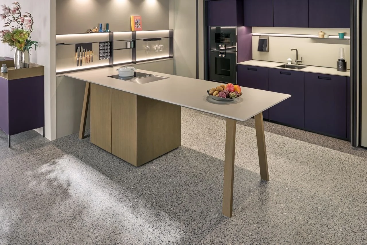 Modern kitchen with grey and purple cabinets, a white countertop island with a bowl of assorted fruits, and a textured grey floor.