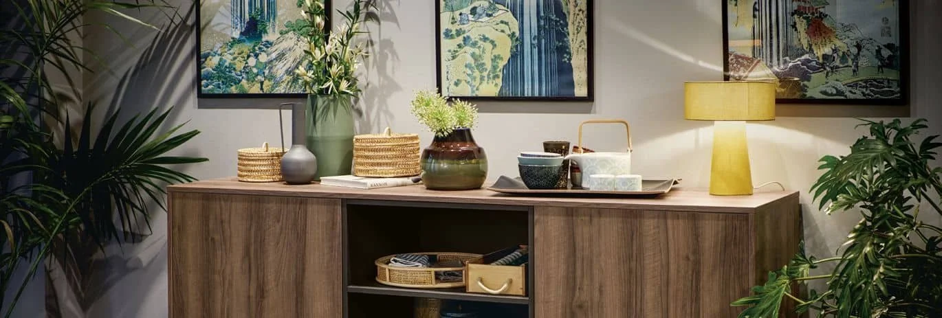 A wooden sideboard with various decorative items including vases, baskets, a lamp, and bowls, with framed artwork and plants in the background.