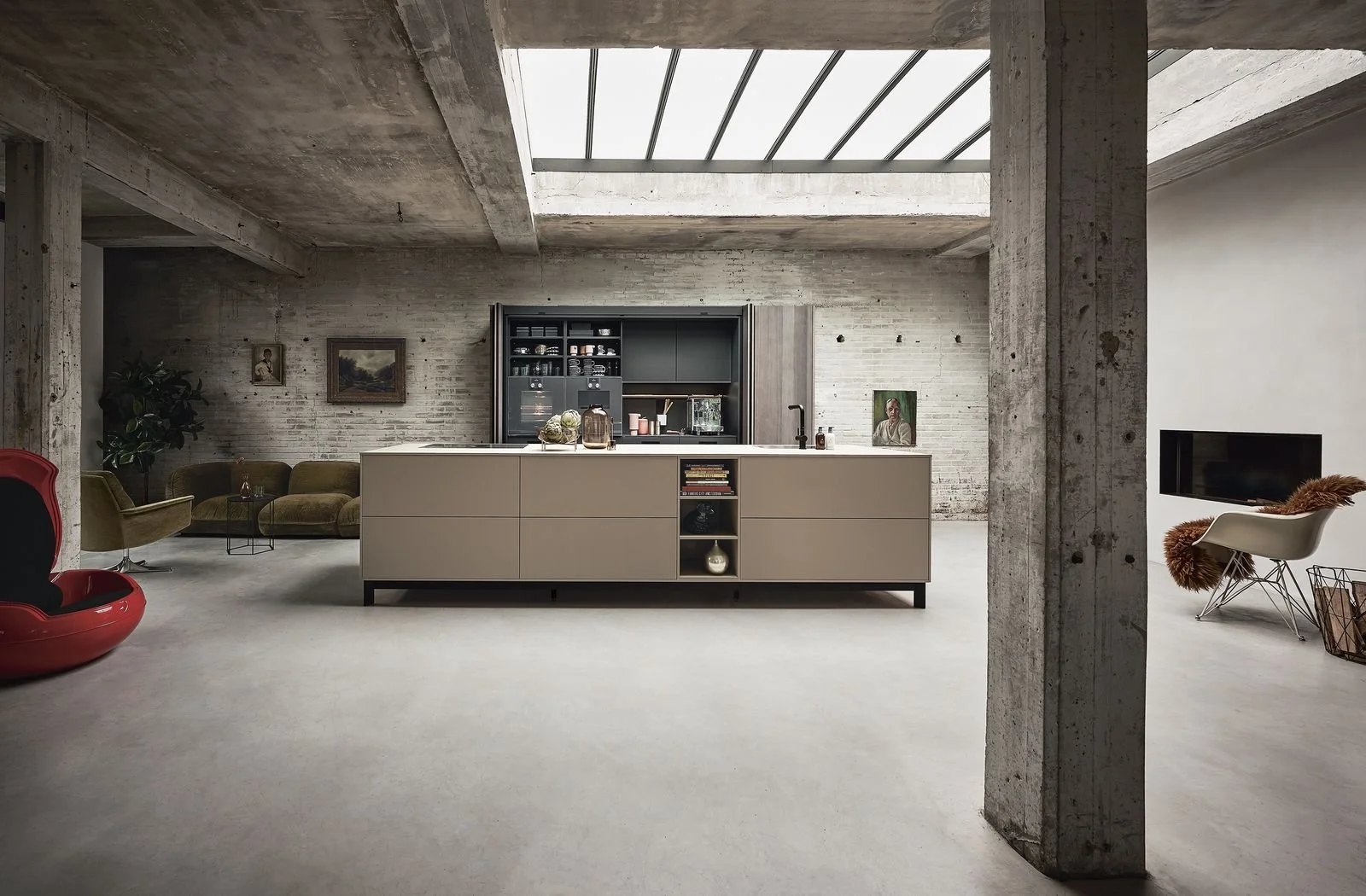 Open plan loft with concrete walls and ceiling, large skylight, modern kitchen with beige cabinetry, living area with olive recliners and a red pod chair, white floor, decorative items, and a wall-mounted TV.