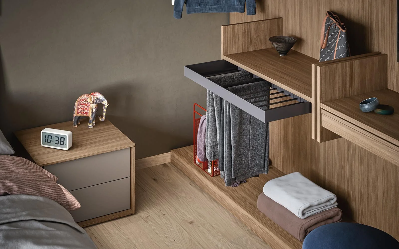Close-up of a bedroom with a wooden nightstand, a clock, decorative elephant, and folded blankets on the floor. There is a wooden storage unit with hanging and stacked clothes, bowls, and a bag on top.