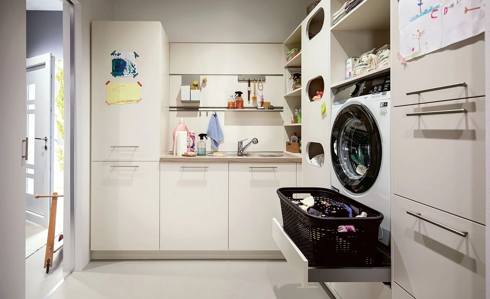 A modern laundry room with white cabinets, a washing machine, a laundry basket, shelves with cleaning supplies, and a small sink.