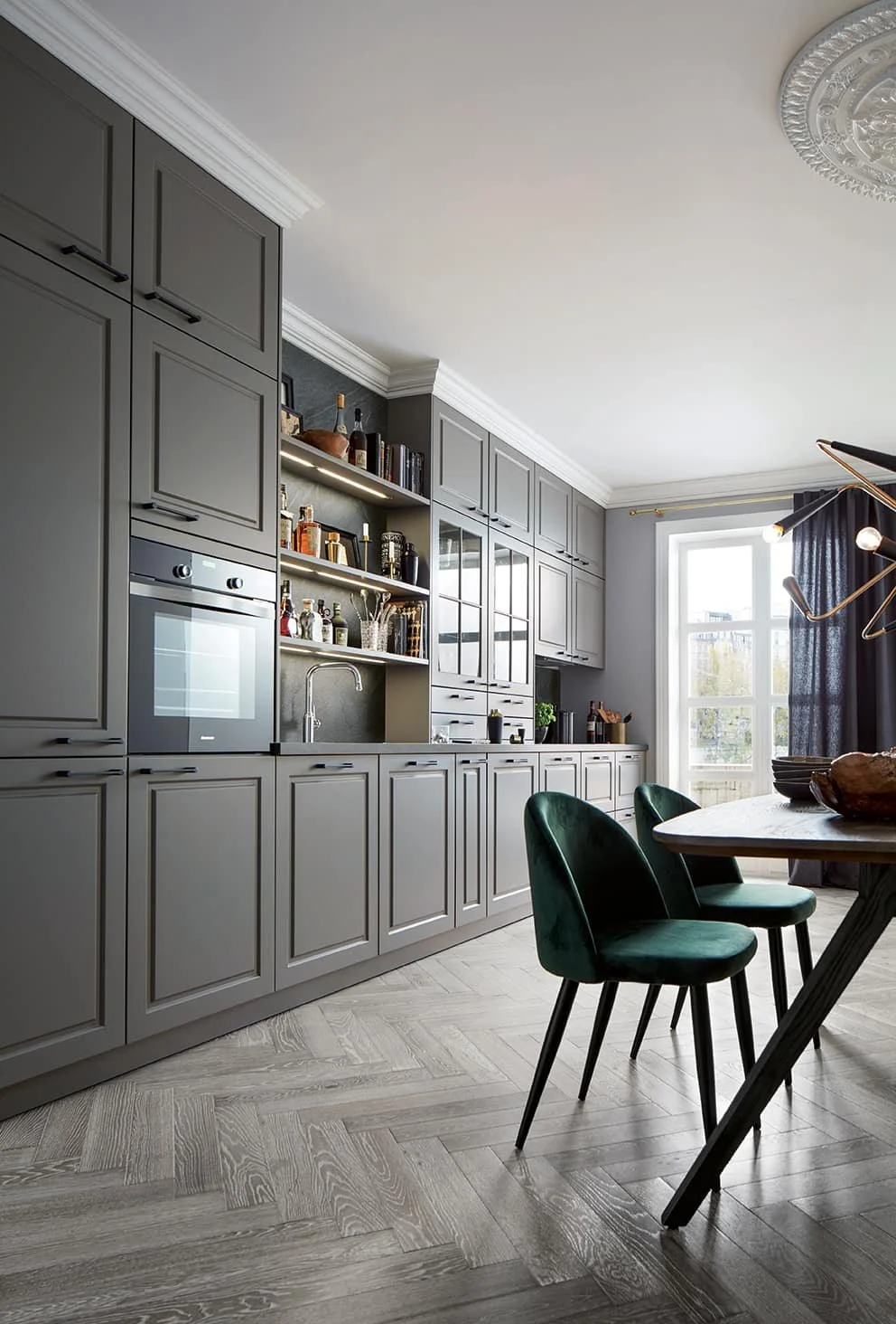 Shaker kitchen with grey cabinets, a wooden dining table, green velvet chairs, and a large window with dark curtains.