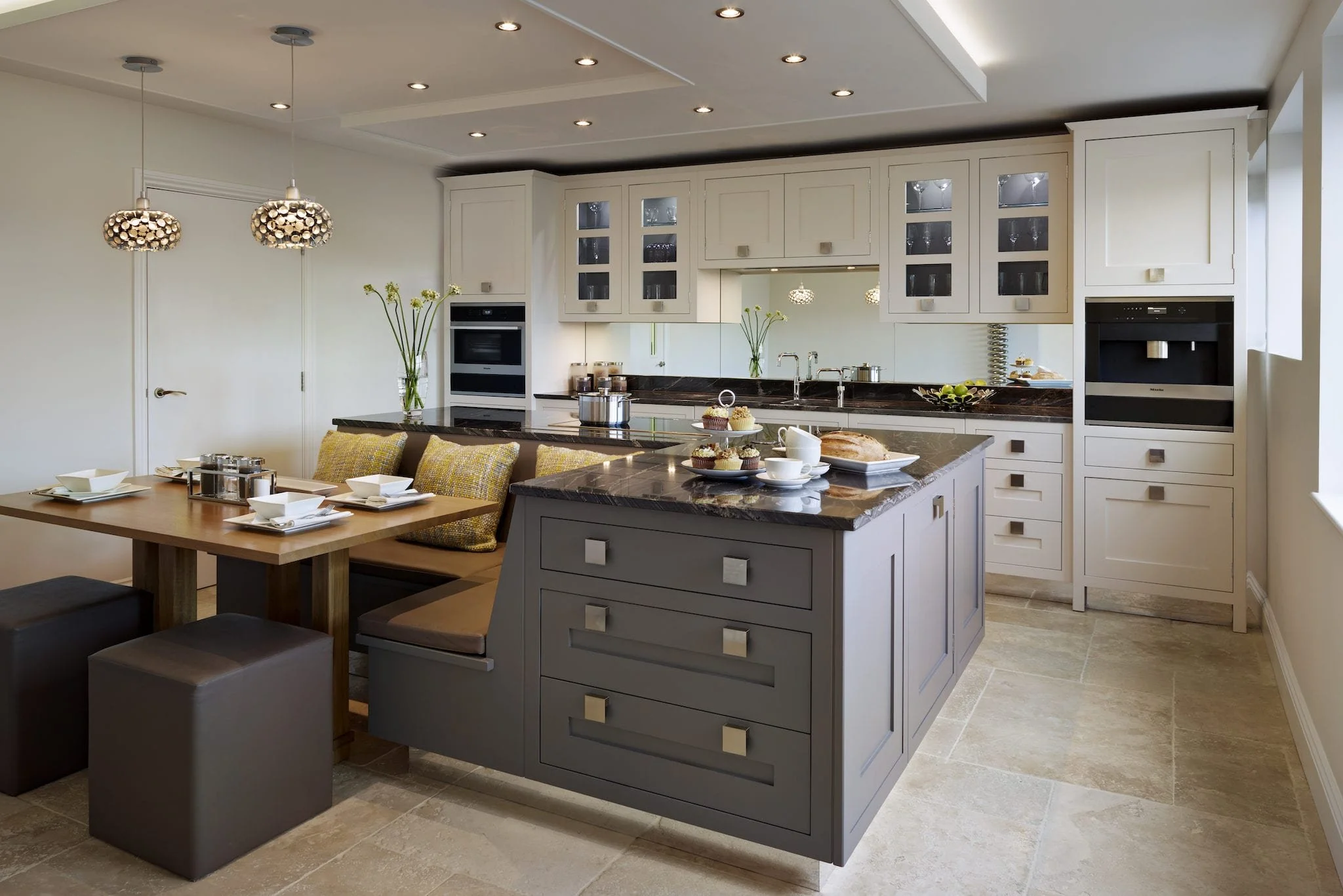 Kitchen with white cabinets, black marble countertops, and a central island with dishes and cups. A dining table with chairs and a bench is set with white dishes, and two pendant lights hang from the ceiling.