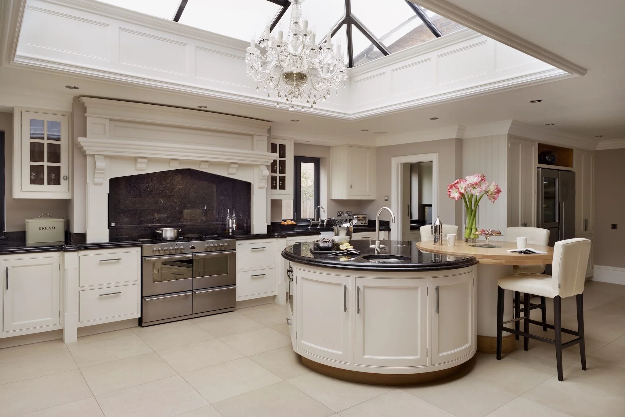 Shaker kitchen with white cabinetry, black countertops, and a large central island. Features a chandelier hanging from a skylight in a high ceiling. There are built-in appliances and a dining area with a vase of pink flowers.