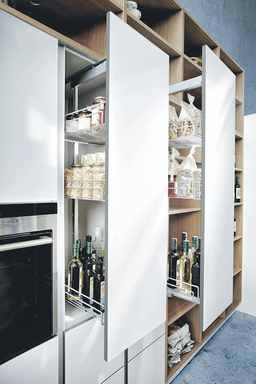 Open kitchen pantry with shelves containing jars, bottles, and food items, next to built-in oven and cabinetry.