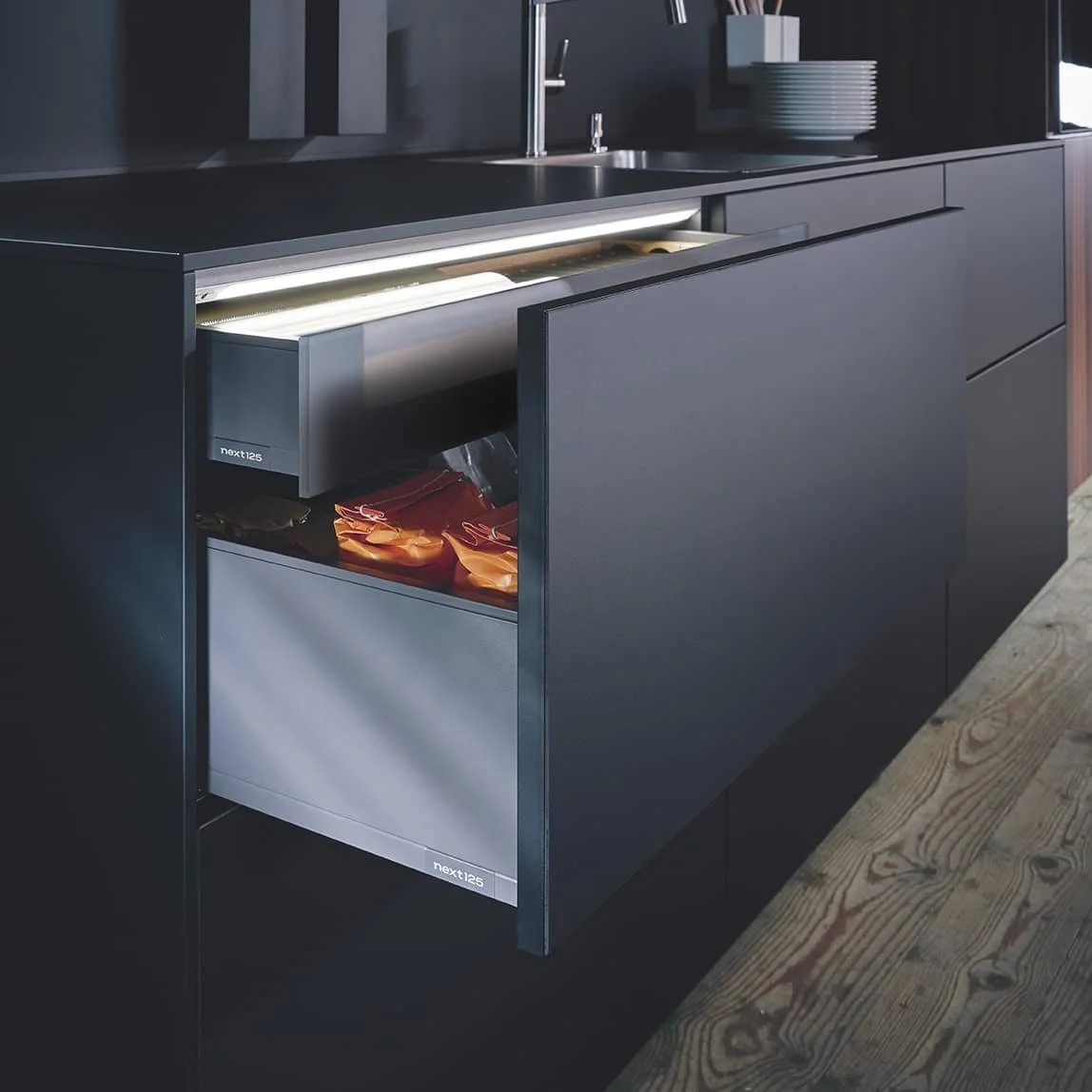 Modern kitchen cabinet with an integrated pull-out trash or recycling drawer, black finish, and wooden flooring.