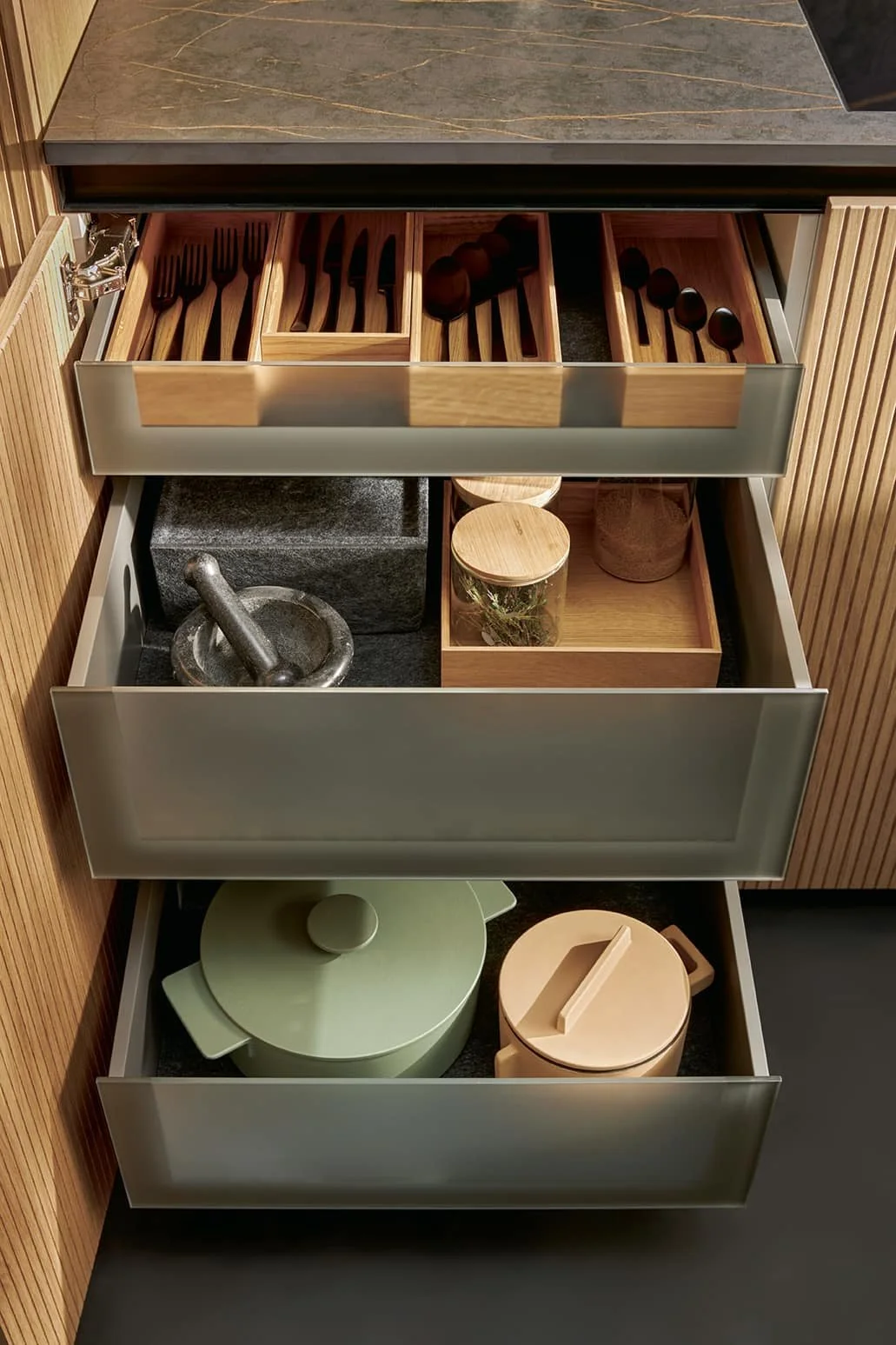 Open kitchen drawer with wooden utensils, a pestle and mortar, jars, and storage containers inside. The kitchen has a dark countertop and wooden cabinetry.
