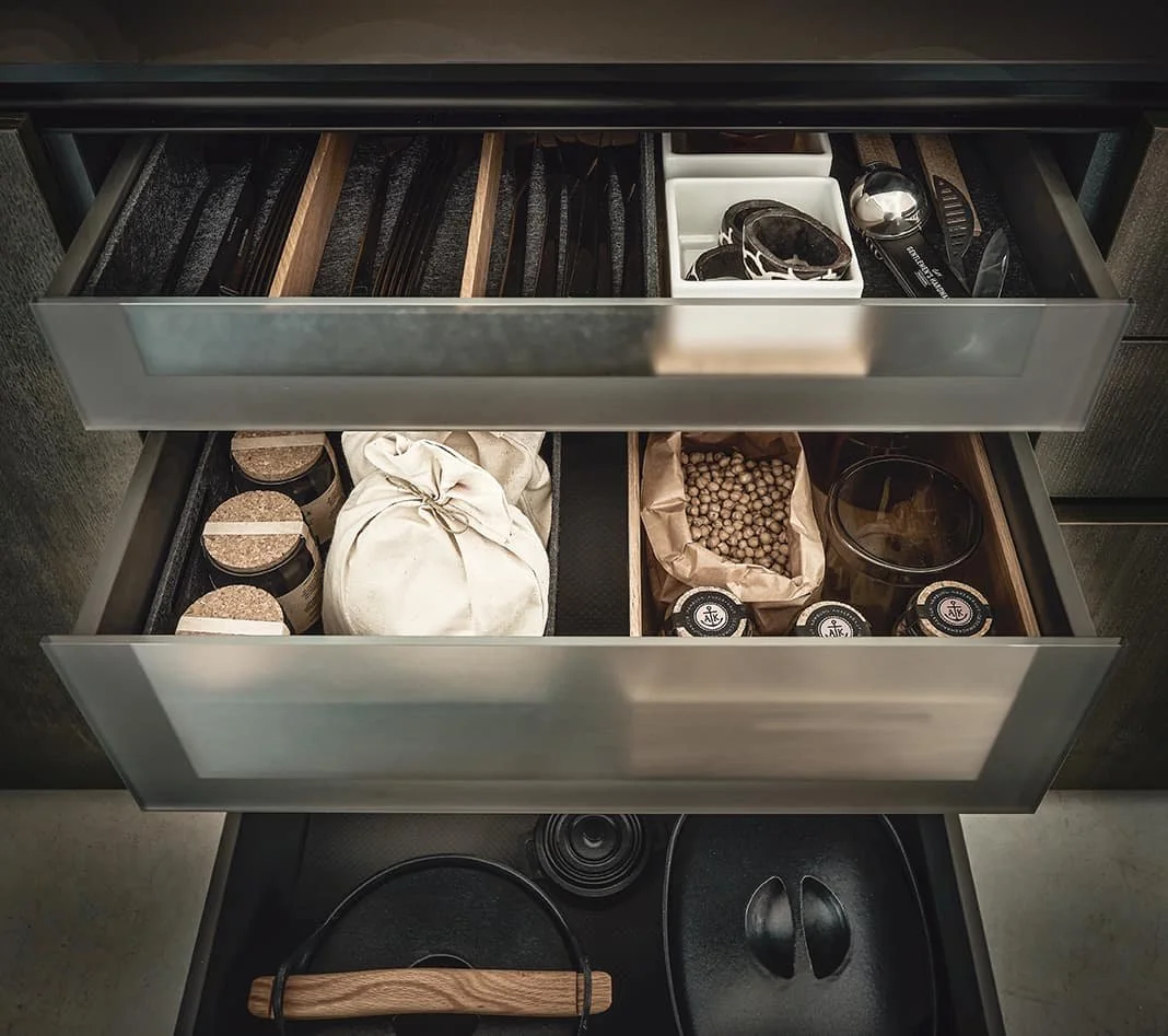 Open kitchen drawers containing spices, jars, a white cloth bag, chickpeas, and jars of other ingredients, with cookware on the stove below.