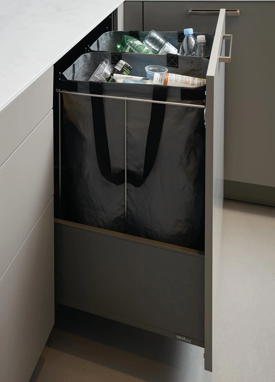 A black reusable bag with handles inside a kitchen drawer, filled with empty plastic bottles and containers for recycling.