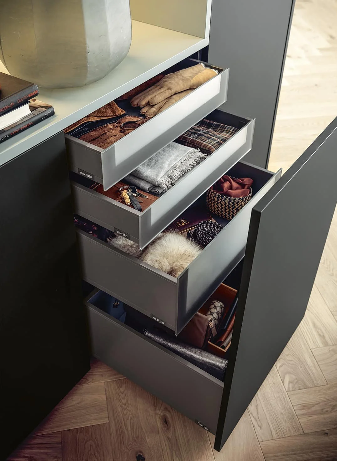 Open grey drawers containing gloves, scarves, keys, a furry item, and miscellaneous accessories, part of a Modern-style cabinet on a wood floor.