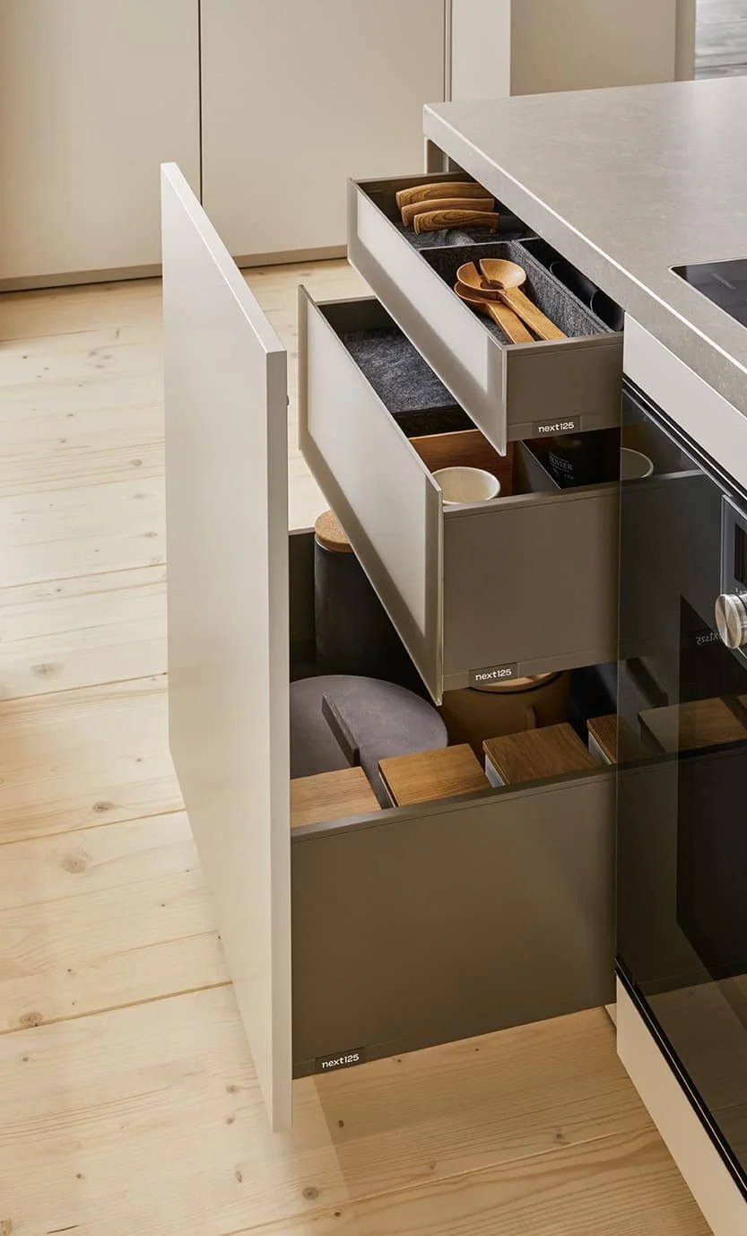 Open kitchen drawer containing wooden spoons and other kitchen utensils, with additional organised drawers below and a light wood floor in the background.