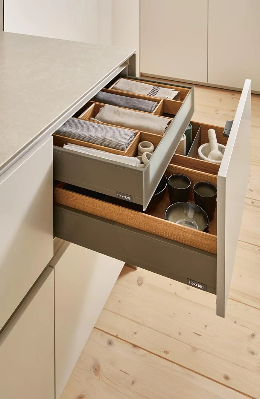 Open kitchen drawer containing neatly folded cloth napkins, small bowls, and cups, with additional dishes stored in the lower section.