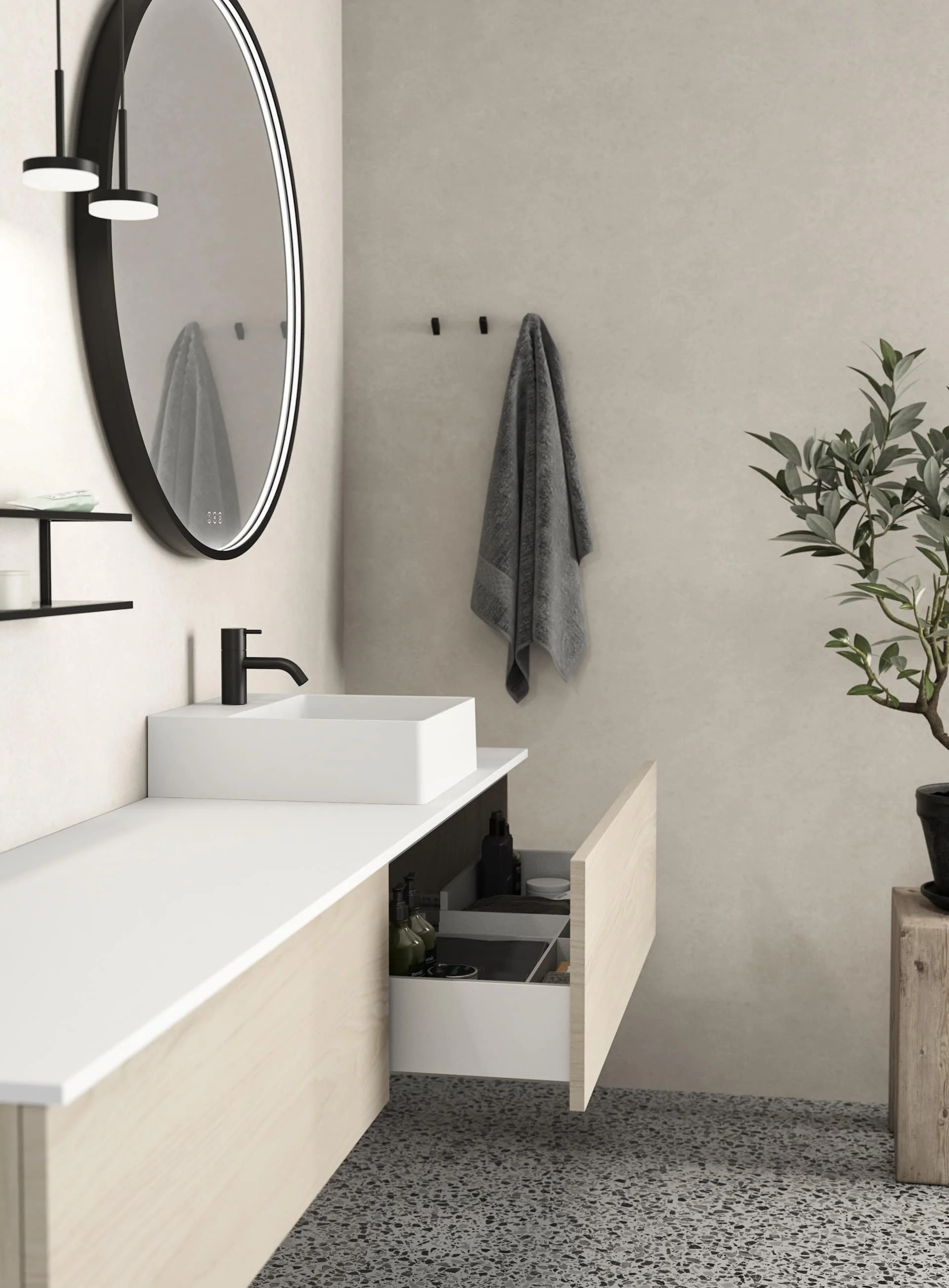 Modern bathroom with light wood vanity, white countertop, vessel sink, black faucet, large oval mirror, black wall-mounted light fixture, potted plant on wooden stand, and terrazzo flooring.