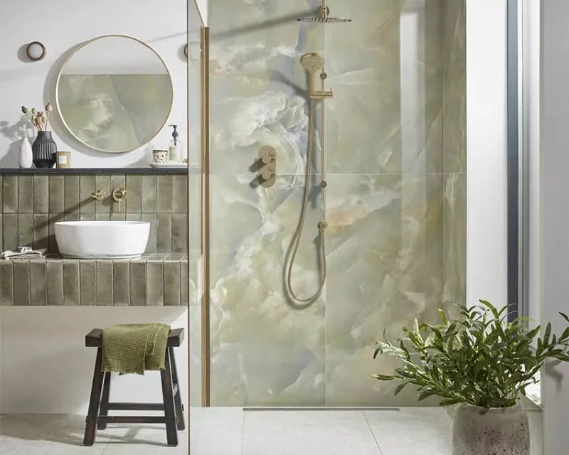 Bathroom with a walk-in shower featuring marble walls, a round mirror above a white vessel sink, a small black stool with a green towel, and a potted plant near a large window.