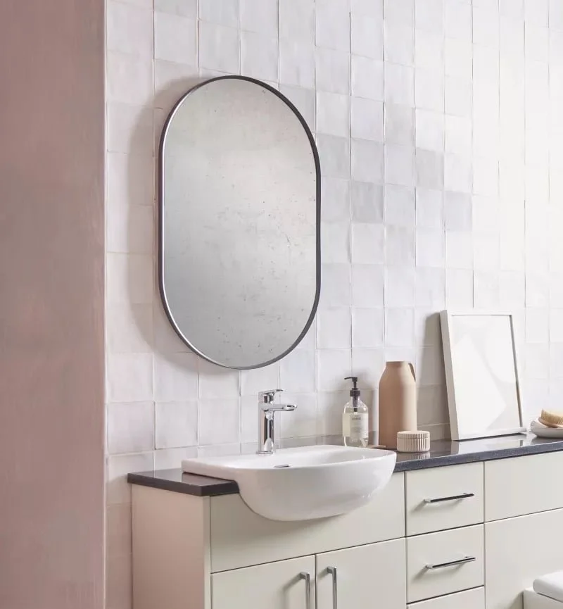 Modern bathroom vanity with a white cabinet, a small white sink, a silver faucet, and a large oval mirror on a tiled wall.