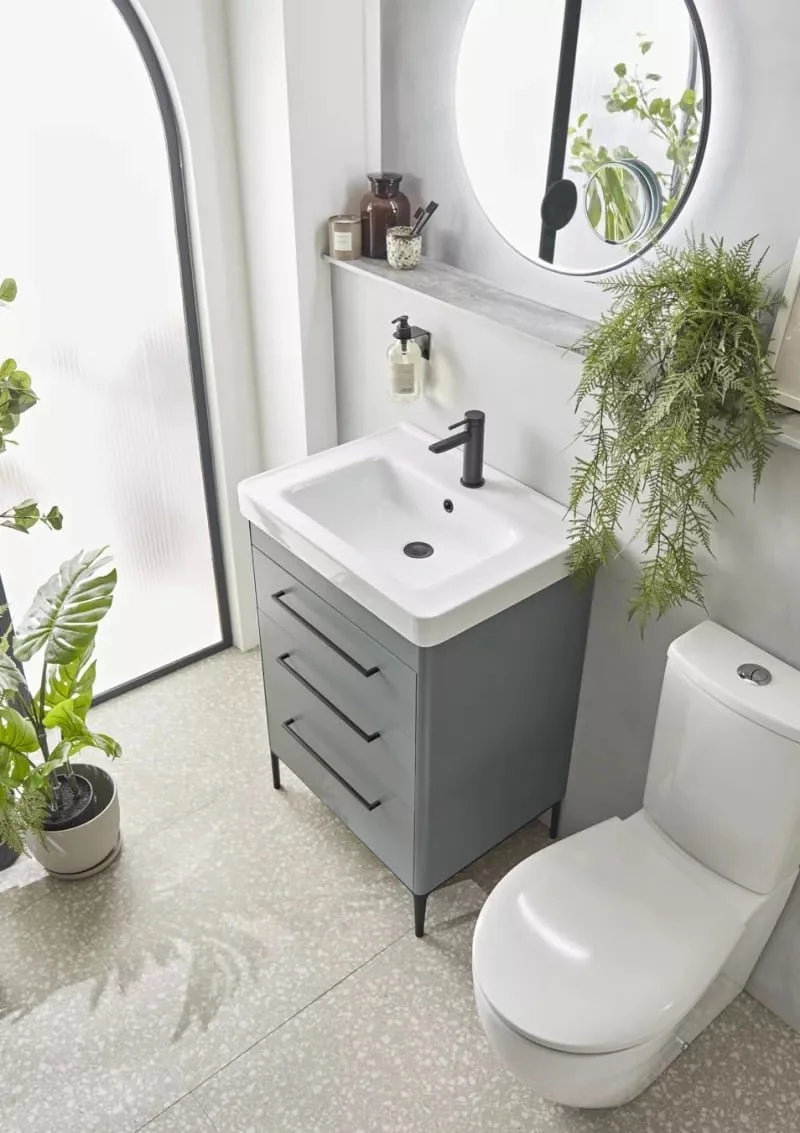Modern bathroom with grey vanity, white sink, black faucet, toilet, round mirror, potted plants, frosted glass door, and white walls.