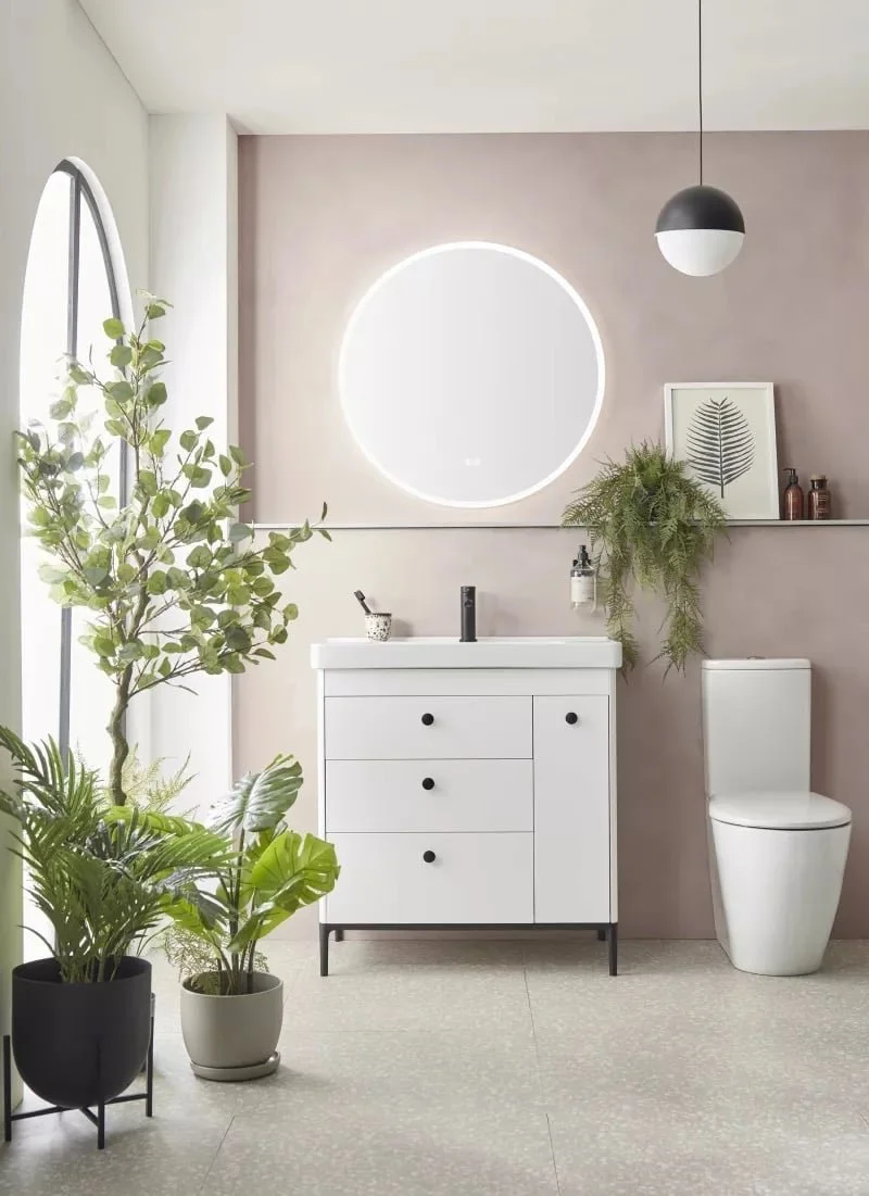 Modern bathroom with white vanity, round illuminated mirror, toilet, green plants, and minimalist decor.
