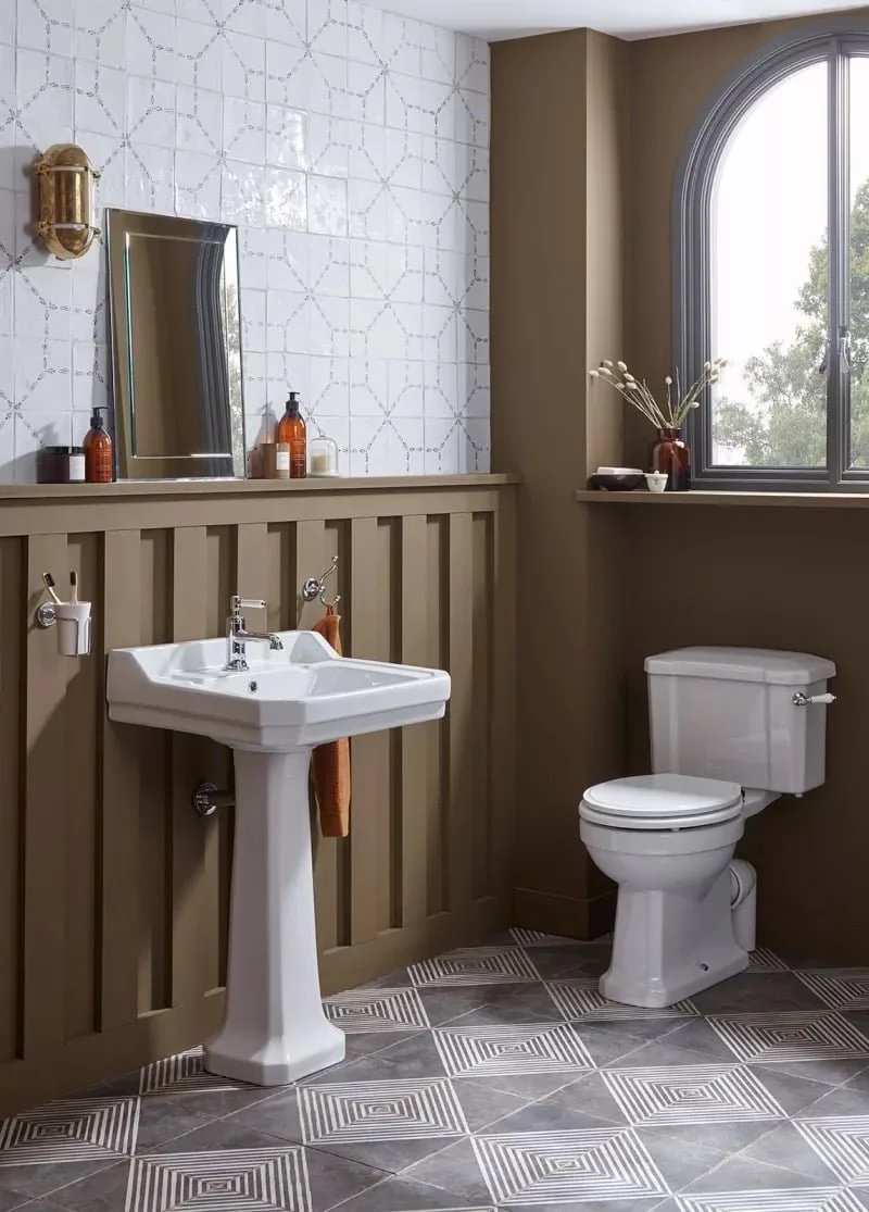 A bathroom with a white pedestal sink, a toilet, a mirror, and a window with outdoor view. Decor includes brown and white tiles, a vase on the window sill, and a light fixture on the wall.