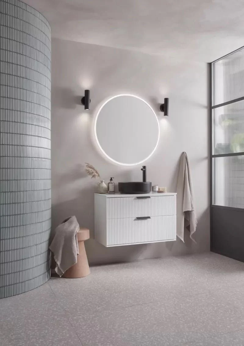 Modern bathroom with a white floating cabinet, a black vessel sink with a matte black faucet, a round backlit mirror, and black wall-mounted light fixtures. There are beige towels hanging, a small beige stool with a towel, and large windows with fros