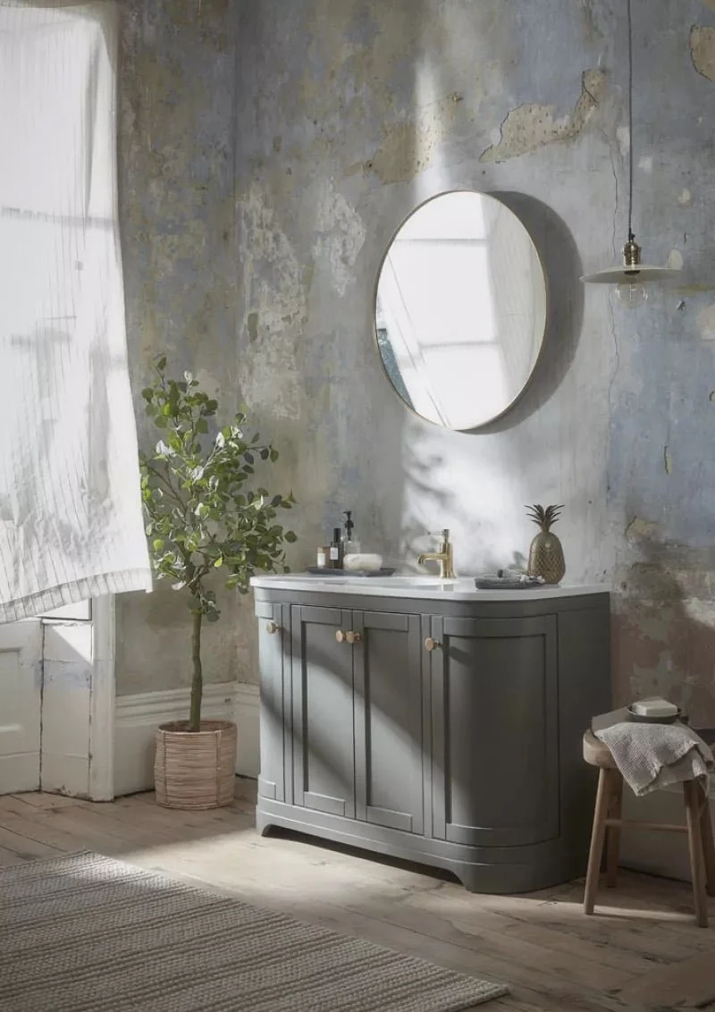 Bathroom with a grey vanity, round mirror, potted plant, pineapple decor, and a stool with a towel, in a rustic space with peeling paint walls and wooden flooring.