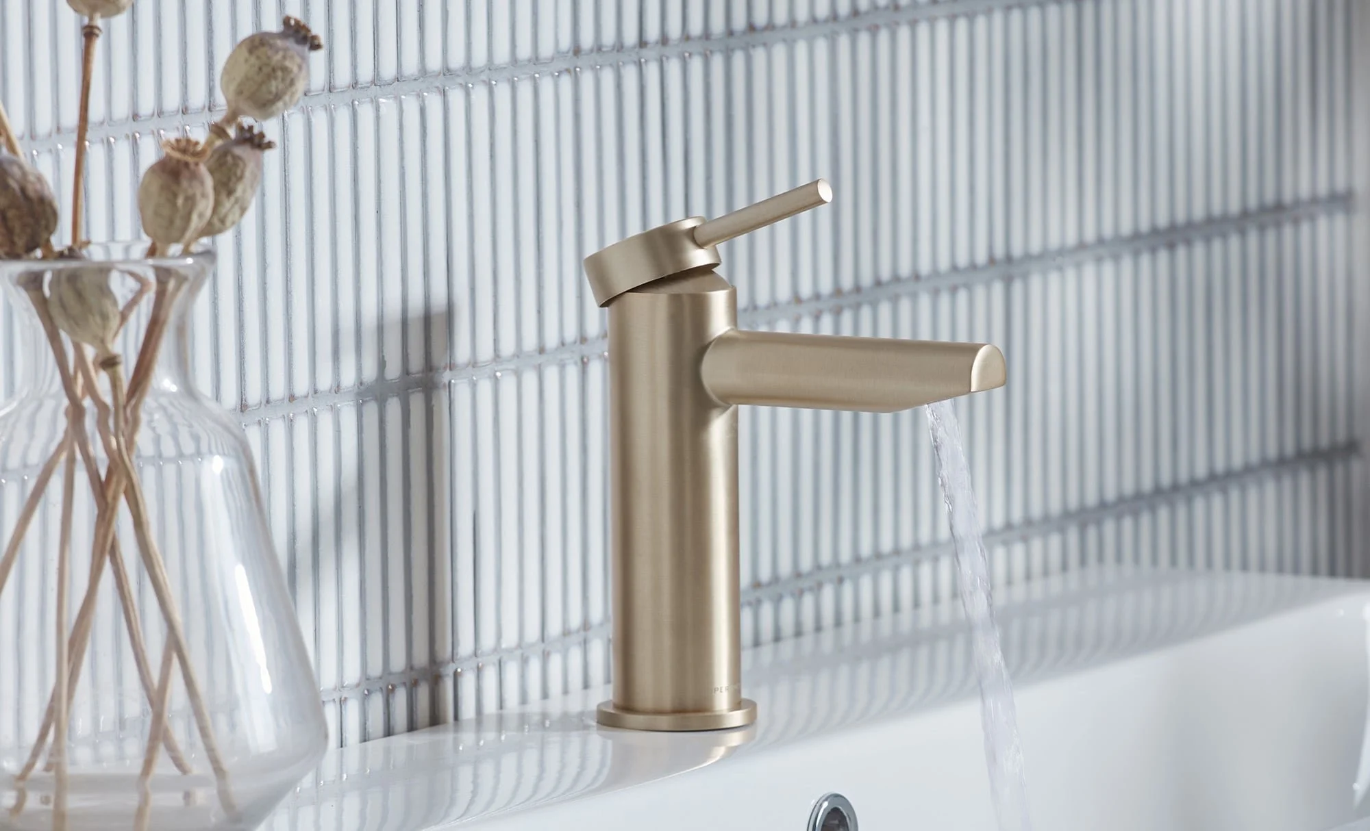 Brushed gold bathroom faucet running water over a white sink, with a clear glass vase containing dried branches nearby, against a white tiled wall.