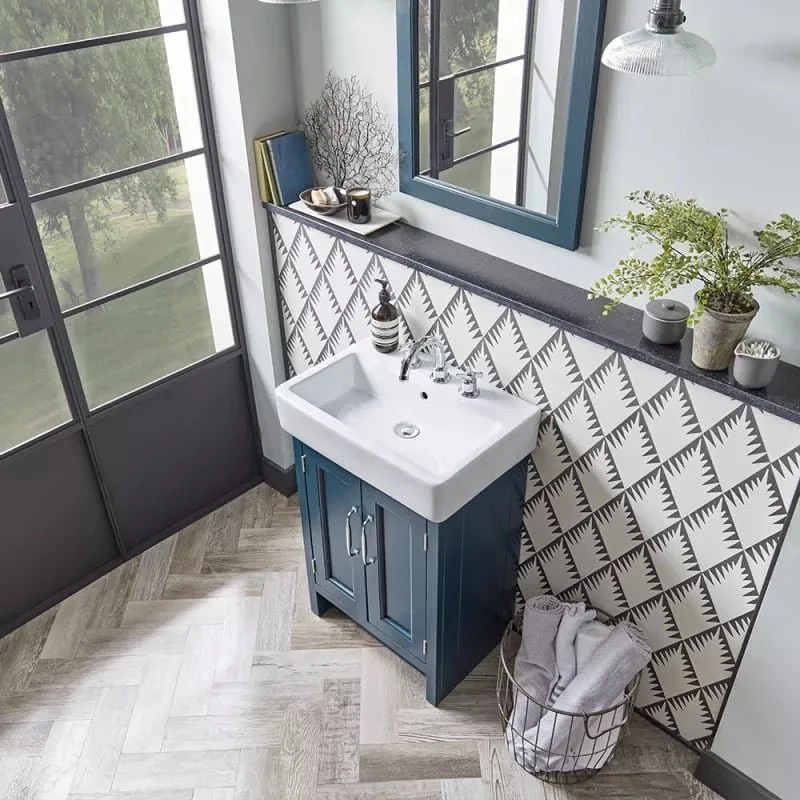 Small bathroom corner with a white sink and blue cabinet, patterned black and white wall, potted plant, mirror, and a window with trees outside.