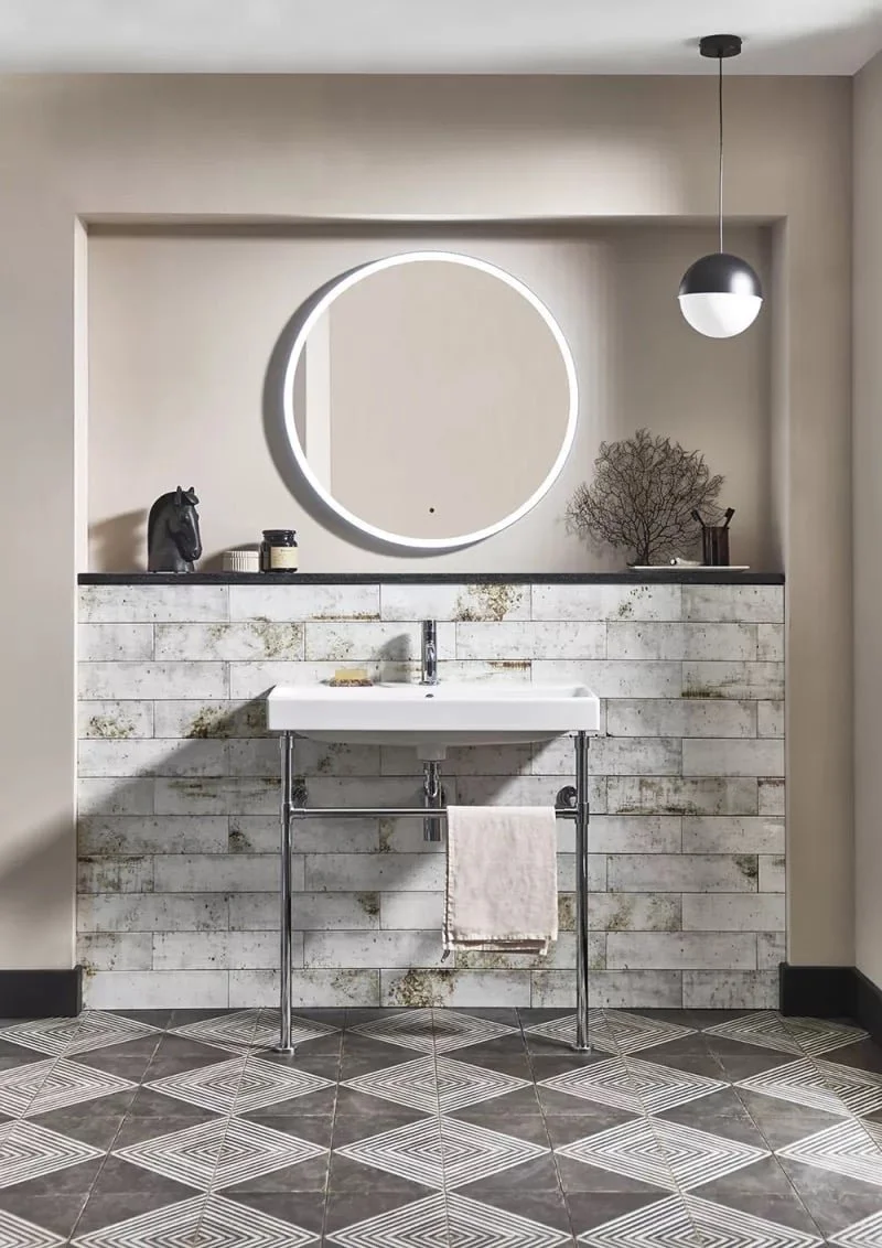 Modern bathroom with white sink, circular mirror with LED lighting, decorative items including a horse bust and a small tree, geometric patterned floor tiles, and hanging pendant light.
