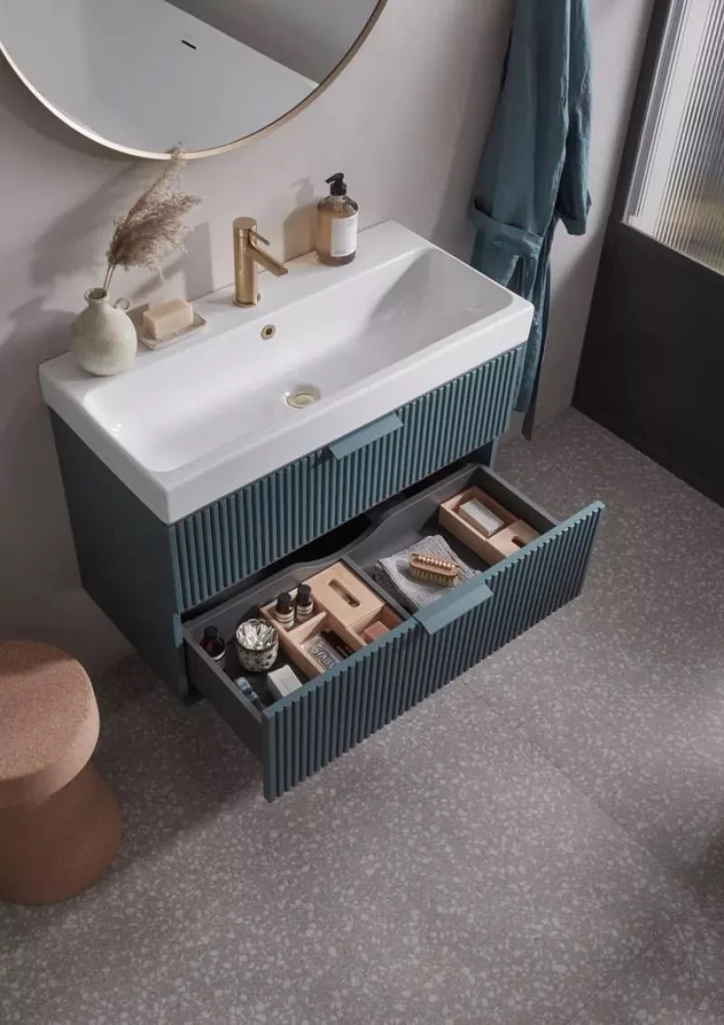 A modern bathroom vanity with a white sink, gold faucet, and an open drawer containing toiletries and grooming products. There is a round mirror above the sink, a beige soap dispenser, and a beige vase with dried pampas grass.
