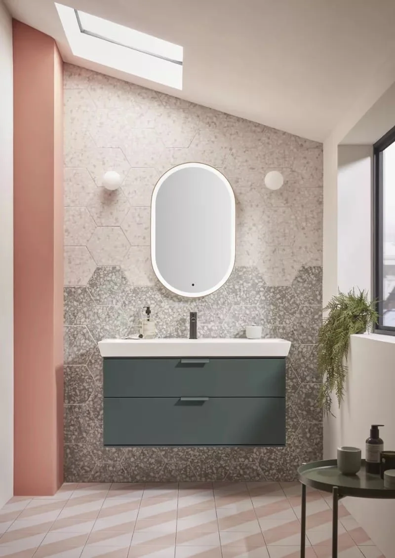 Modern bathroom with a floating teal vanity, an oval mirror, hexagonal wall tiles, a skylight, a window with black frame, and a small table with toiletries.