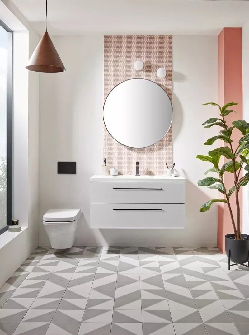 Modern bathroom with a white floating vanity, round mirror, two spherical wall lights, potted plant, patterned floor tiles, and a pink accent wall.
