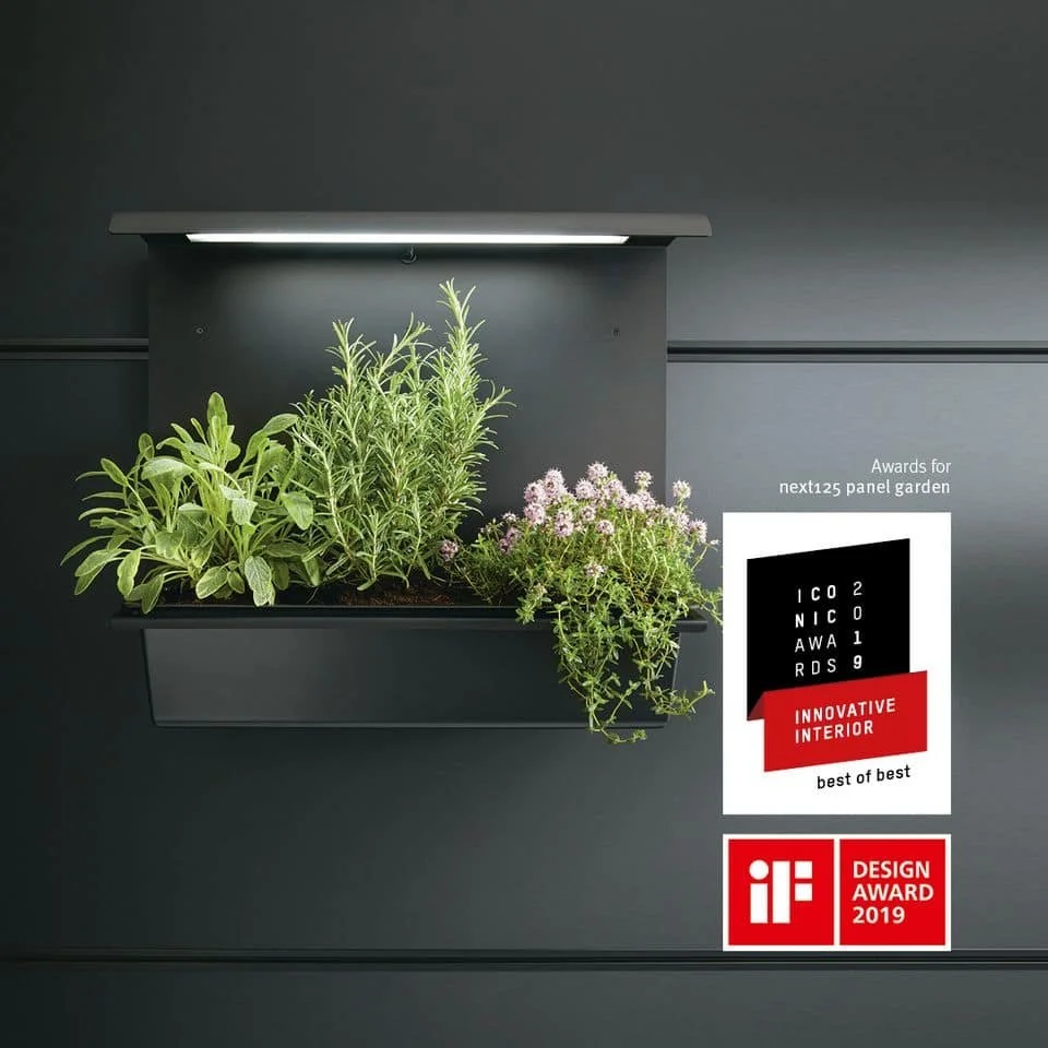 Indoor plant panel garden with green herbs and flowering plants under a light, mounted on a dark gray wall. It features awards for innovative interior design, including the iF Design Award 2019.