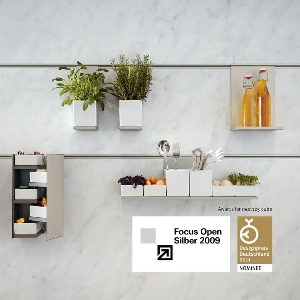 Minimalist kitchen shelf with potted herbs, jars with beverages, storage boxes with vegetables, and utensils.