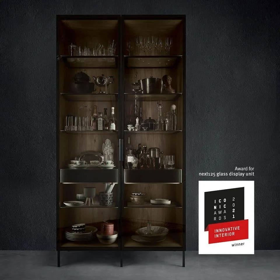 A tall wooden display cabinet with glass doors, housing various glasses, bottles, and decorative items, against a dark textured wall, with award signage on the right side.