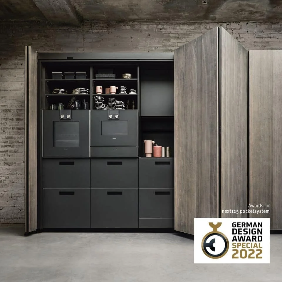 Modern kitchen cabinet with open wooden doors revealing black cabinetry and pink and black dishes, next to a textured grey brick wall.