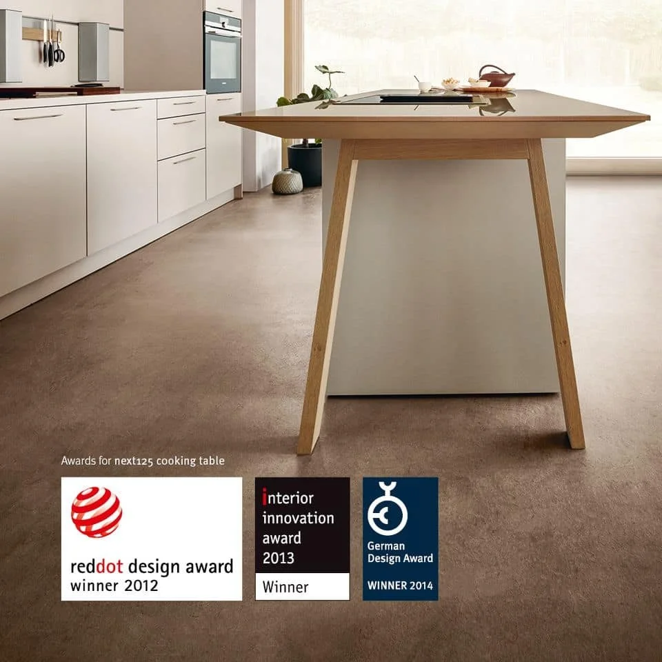 Modern kitchen with white cabinetry and a wooden cooking table in the center, decorated with awards at the bottom left.