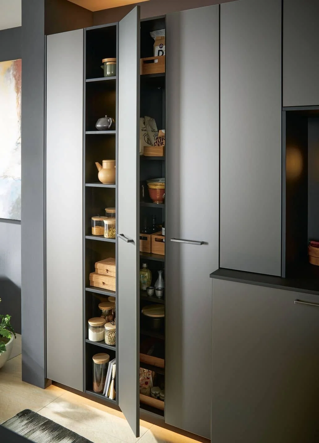 A modern kitchen cabinet with grey doors, some open revealing shelves filled with jars, bowls, boxes, and bottles.